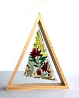 A triangular wooden frame containing a fused glass triangle depicting green leaves, red flowers, and yellow blossoms inspired by Australian native blooms.