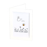 A white greeting card with a folded-over back, featuring the words 'Bee HAPPY' in black script and flower line drawings near the bottom, plus a small gold-and-black bee charm attached.